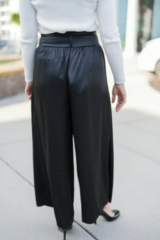 Back view of the Black Satin Wide-Leg Trousers showcasing the subtle elasticated back waistband and smooth, flowy back silhouette. Paired with a fitted off-white ribbed long-sleeve top tucked in and black stiletto heels.