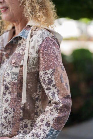 Side-profile portrait highlighting the sleeve and shoulder construction of a vintage-inspired patchwork hooded jacket. Displays the contrast cream drawstring cord, pointed collar design, chest flap pocket with copper hardware, and multi-patterned paisley and botanical fabric panels.
