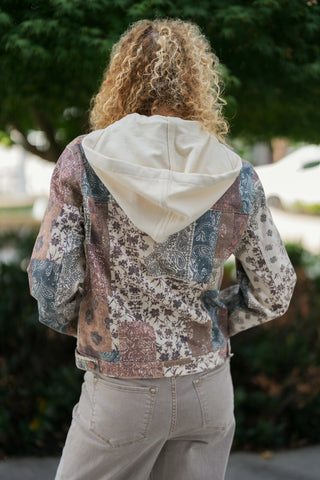 Back view of a woman wearing a patchwork utility jacket, showcasing the integrated soft cream jersey hood draped over the upper back. Highlights the back yoke design, structured waist seam, and continuous multi-pattern patchwork print paired with light taupe jeans.