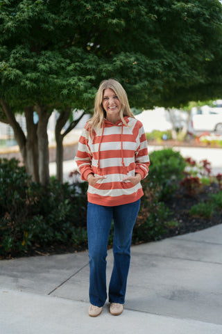 Woman standing outdoors in a rust-orange and cream broad horizontal striped pullover hoodie featuring a front kangaroo pocket, braided drawstring, and relaxed raglan sleeves. Styled casually with classic blue denim jeans and light-colored sneakers against a lush green garden background.