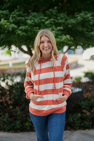 Mid-shot of a smiling woman posing in a rust-orange and off-white striped hoodie with hands tucked into the front kangaroo pouch. Highlights the cozy, casual fit, ribbed cuffs, and textured twisted drawstrings paired with blue jeans and subtle gold bracelets.