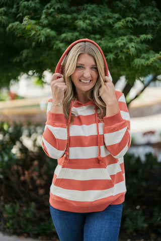 Close-up portrait view of a woman holding the hood up on a rust-orange and cream striped pullover hoodie, showcasing the hood design, braided cords with knot details, and the soft, comfortable fabric texture.
