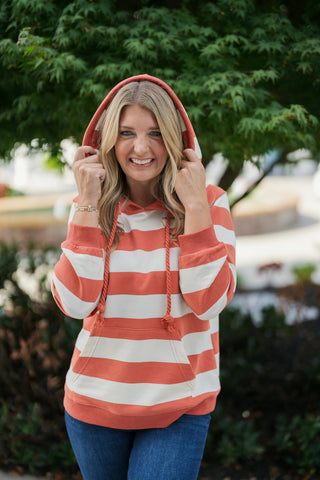Close-up view of a smiling woman frame-holding the hood of a rust-orange and off-white striped hoodie over her head, highlighting the relaxed neck construction, drawstring accents, and warm autumn color palette