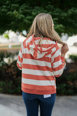 Back view of a blonde woman wearing a rust-orange and cream broad-striped pullover hoodie and blue denim jeans. Showcases the back profile of the attached hood, ribbed hem finish, relaxed fit, and continuous horizontal stripe pattern across the back.