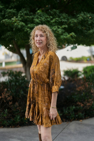 Three-quarter view of a woman in an mustard-gold vintage floral satin mini dress, showcasing the relaxed bodice fit, adjustable tie waist, and voluminous elbow-length sleeves with structured button cuffs.