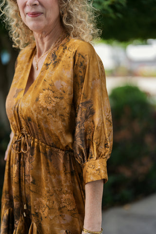Close-up side-angle shot highlighting the luxurious satin sheen and dark brown botanical print of the ochre mini dress. Focuses on the deep V-neckline, fine gathering at the cinched waist, and cuffed blouson sleeve detail