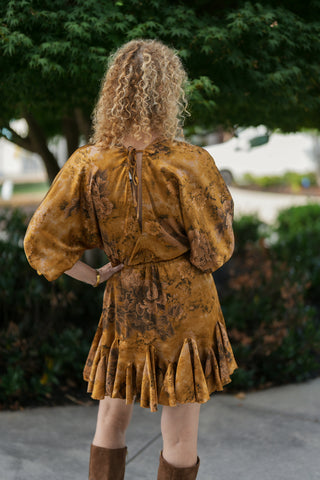Back view of a woman wearing an ochre floral satin drop-waist dress. Highlights the keyhole back closure with slender neck ties, subtle gathering at the elasticated waist, and the full movement of the pleated flounce hemline.
