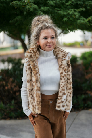 Three-quarter view of a smiling woman wearing an open-front faux fur leopard print vest featuring a plush lapel collar. Highlights the rich animal print pattern, cropped waist length, and contrast against an off-white fitted ribbed long-sleeve turtleneck.