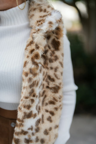 Extreme close-up shot focusing on the soft, tactile texture of the faux fur leopard print vest. Showcases the realistic spotted pattern in tan, brown, and ivory tones paired with the clean vertical knit lines of a white turtleneck.