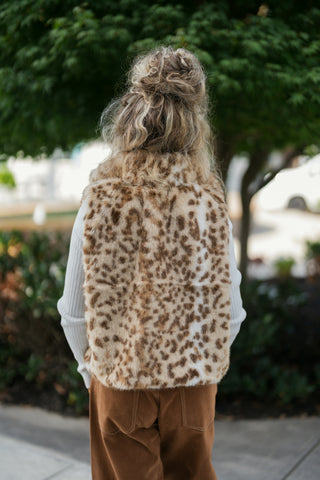 Back view of a woman wearing the leopard print faux fur vest, showing the seamless pattern layout and straight hemline across the back. Paired over a long-sleeve ribbed white top and brown suede pants.