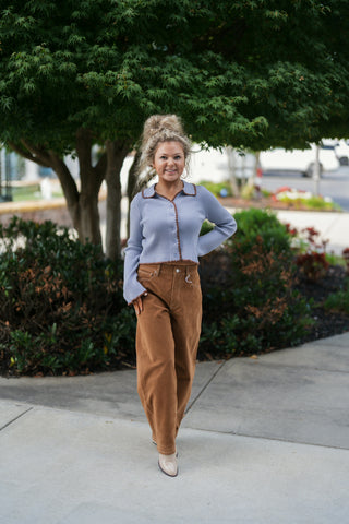 Woman posing outdoors in a periwinkle blue ribbed knit cardigan sweater featuring contrasting brown scalloped ric-rac trim along the collar, button placket, hem, and sleeve cuffs. Styled casually with high-waisted camel brown suede wide-leg trousers, pointed-toe boots, gold hoop earrings, and a high curly updo against a leafy green tree background.