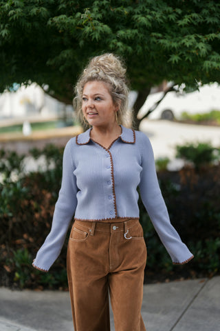 Three-quarter portrait view of a woman wearing a periwinkle blue button-down ribbed cardigan with a flat spread collar. Highlights the soft, fitted silhouette, tonal front buttons, subtle flare at the cuffs, and eye-catching brown embroidered scalloped edging