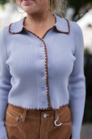Close-up shot highlighting the texture and construction of a lavender-blue ribbed knit cardigan. Focuses on the matching periwinkle buttons, fine ribbed fabric weave, spread collar structure, and detailed brown scalloped trim along the front opening and cropped hemline.