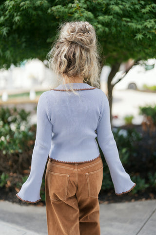 Back view of a woman wearing the periwinkle blue ribbed cardigan, showcasing the clean vertical ribbing, fitted back cut, slightly flared long sleeves, and brown scalloped trim lining the collar, hem, and cuffs. Paired with camel suede pants.