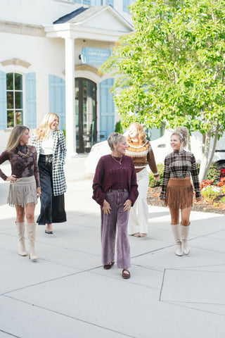 Full-length group shot of five women laughing and walking outdoors, showcasing cohesive autumn style combinations. Highlights a mix of seasonal textures including fine corduroy, suede fringe, knit crochet, Fair Isle patterns, and tailored houndstooth outerwear in shades of burgundy, plum, chocolate brown, cream, and taupe. Photographed on a sunlit plaza in front of modern architecture and lush greenery.