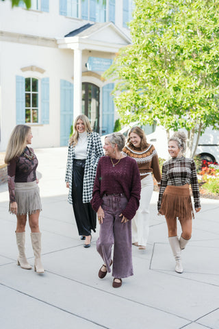 Five women walking together outdoors in coordinated fall fashion outfits featuring rich earth tones, textures, and patterns. Outfits include a sheer dark floral top with a beige fringe mini skirt and tall boots; a black and white houndstooth coat layered over a white turtleneck and black wide-leg trousers; a deep burgundy crochet knit sweater paired with plum corduroy scalloped-pocket pants and brown Mary Janes; a brown pumpkin Fair Isle sweater with cream trousers; and a brown velvet-textured plaid mock ne