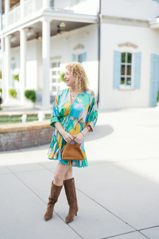 Woman posing outdoors in a colorful abstract floral mini dress featuring a vibrant print in teal blue, mustard yellow, sage green, and soft blush pink. The dress has a flattering V-neckline, elbow-length balloon sleeves with wide cuffs, an elasticized drawstring tie waist, and a playful ruffled drop-hem skirt. Styled with tall brown suede knee-high boots and a matching suede handbag in front of a modern white building with light blue shutters.