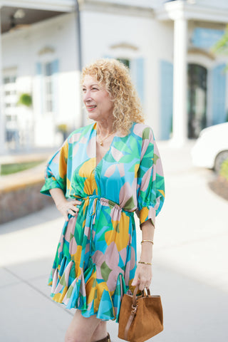 A three-quarter view highlighting the relaxed silhouette and abstract botanical pattern of the mini dress. Features include a deep V-neckline, an adjustable drawstring waist with tassel ties, three-quarter length volume sleeves, and a tiered ruffle hem. Paired with fine gold jewelry and a brown suede handbag for an elevated transitional outfit.