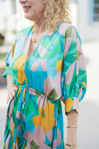 Close-up view emphasizing the intricate fabric pattern and construction details of the V-neck mini dress. Shows the cinched gather waistline with matching tassel drawstrings, structured elbow-length balloon sleeves, and smooth silk-like texture in bright teal, yellow, green, and blush tones, styled with delicate gold bangles and a dainty pendant necklace.