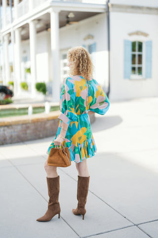 Back view showcasing the flowing silhouette and vibrant rear pattern of the abstract printed mini dress. Details include a keyhole back closure with a small tie, relaxed elbow-length sleeves, an elastic waist gather, and a flouncy ruffled skirt hem. Styled with brown suede tall boots and a suede mini tote against a bright outdoor plaza backdrop.