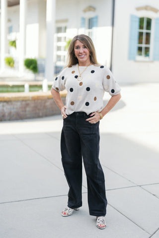 Woman posing outdoors in a beige short-sleeve knit sweater adorned with multi-colored polka dots in black, taupe, and bronze tones. The crewneck top features raglan sleeves with ribbed cuffs and is casually tucked into high-waisted black wide-leg jeans paired with open-toe sandals. Styled with a gold chain link oval pendant necklace and stacked gold bracelets against an upscale building courtyard.