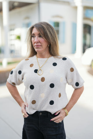 A portrait view of the Polka Dot Short-Sleeve Knit Sweater highlighting the soft knit texture and ribbed crew neckline. The oatmeal-beige background is accented with playful polka dots in dark and neutral shades, complemented by a statement gold link necklace and chic raglan short sleeves for an elevated casual look.