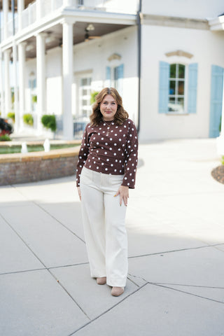 Woman posing outdoors in a chocolate brown long-sleeve top with white polka dots, styled with high-waisted cream corduroy wide-leg pants and taupe suede shoes. The top features a crew neckline, relaxed drop-shoulder long sleeves, and a tailored waist panel. Styled in front of a modern white building with light blue shutters.