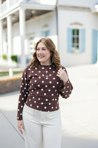 A portrait view highlighting the detail of the brown and white polka dot crewneck top, styled with a gold pendant cross necklace and small hoop earrings. The long-sleeve top shows drop-shoulder seams, bell-style cuffs, and a defined hemband. Paired with cream high-waisted corduroy pants featuring front patch pockets.