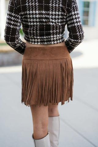 Back view from the waist down focusing on the rear coverage and straight hemline of the chestnut faux-suede fringe skirt. Highlights the cropped lower hem of the brown plaid top, smooth waistband seam, and tall off-white boots.