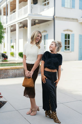 Two women posing outdoors together in elevated autumn outfits. On the right, a woman wears the black satin lace-trim midi skirt with a black fuzzy cropped short-sleeve sweater and leopard print knee-high boots. On the left, a woman pairs an off-white fuzzy short-sleeve sweater with a chocolate brown lace-hem midi skirt, nude braided heel sandals, and a tan suede handbag in front of a white building