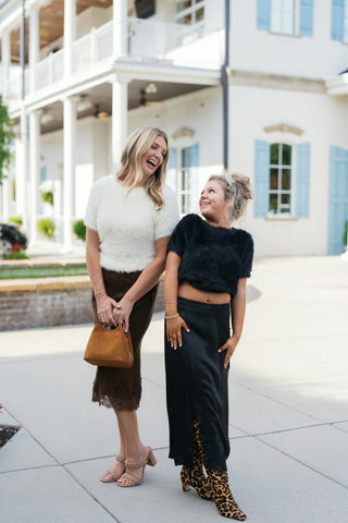 Two women posing outdoors showcasing both style variations: one wearing the off-white plush short-sleeve sweater tucked into a brown satin lace skirt, and the other wearing the black cropped version with a black satin midi skirt and leopard print boots against a white building background.