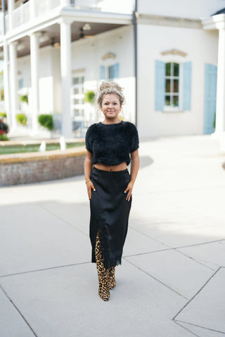 Full-length front view of a woman wearing a black satin midi skirt featuring delicate black lace appliques at the hip and an asymmetrical front slit trimmed in black eyelash lace. Styled with a cropped black fuzzy short-sleeve sweater, gold hoop earrings, and statement leopard print pointed-toe boots outdoors.