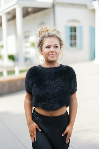 Portrait shot of a woman in a black cropped eyelash knit short-sleeve sweater with a crew neckline. Showcases the textured plush material paired with gold hoop earrings and a high curly updo.   