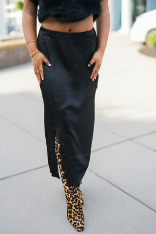 Mid-section shot highlighting the waist and front detail of a black satin midi skirt with intricate black lace inset panels along the upper hip and lower slit. Paired with a cropped black fuzzy top and bold leopard print boots, accentuating the high-waisted fluid silhouette.