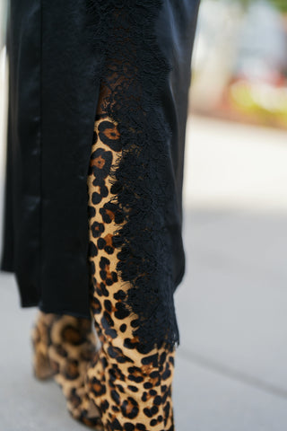 Extreme close-up view of the side slit on a black satin midi skirt, showcasing the delicate floral eyelash lace trim against a statement leopard print leather boot. Illustrates the high-contrast texture and fine lacework construction.