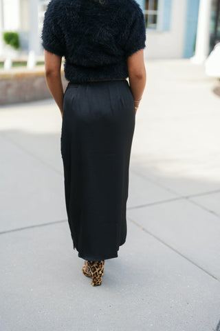 Back view of a woman wearing a sleek black satin midi skirt paired with a cropped black plush fuzzy sweater and leopard print block-heel boots. Highlights the smooth, tailored back fit and clean hemline as she walks outdoors.