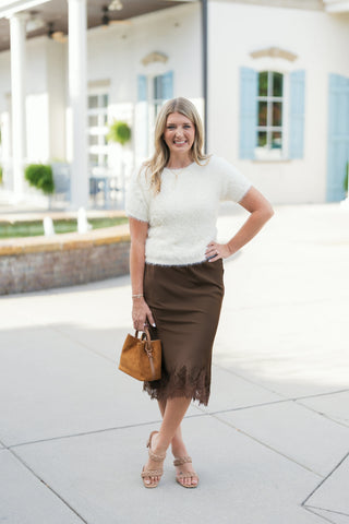 Woman standing outdoors in an off-white plush eyelash knit short-sleeve sweater featuring a crew neck and ribbed hem. Styled with a dark brown lace-trim satin midi skirt, braided block-heel sandals, and a small tan suede handbag.