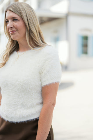 Three-quarter portrait of a smiling woman posing in an off-white fluffy eyelash knit short-sleeve sweater, showing the textured fabric, neat crew neckline, and fine gold necklace detail.