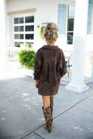 Back view of a woman wearing a matching chocolate brown faux suede set, showcasing the relaxed fit and back yoke detail of the jacket over a faux suede mini skirt. Styled with eye-catching knee-high leopard print boots on a patio setting.