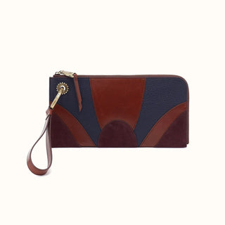 Front view of a rectangular, color-blocked leather wristlet wallet against a solid white background. The mixed-media design features an Art Deco-inspired geometric arch pattern crafted from navy blue pebbled leather, smooth cognac brown leather, and deep burgundy suede. A detachable cognac leather wrist strap connects to the left side via an antique brass lobster clasp and a distinct sunburst-shaped decorative hardware piece. The top zipper closure is finished with a matching smooth leather zipper pull.