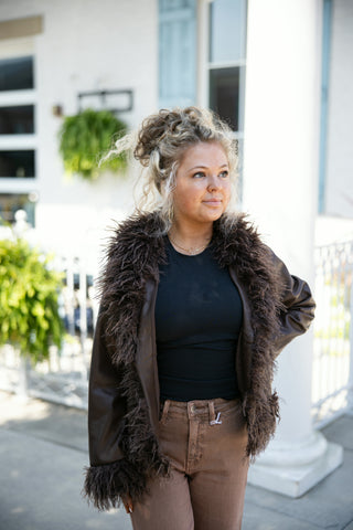 A young woman with curly blonde hair styled in a high bun posing outdoors in a dark brown leather jacket trimmed with shaggy, feather-like faux fur along the lapels, open front, and sleeve cuffs. The jacket is layered over a simple black fitted crewneck top and paired with medium-brown denim jeans. She wears a delicate gold nameplate necklace and stands with one hand on her hip against a sunlit white building backdrop.