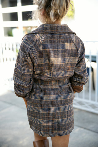 Back view of the matching plaid two-piece set, featuring the structured back yoke and raglan seam details of the short-sleeved trench jacket fastened at the waist with a self-tie belt over a fitted plaid mini skirt. Styled with a messy curly updo on a concrete outdoor walkway.