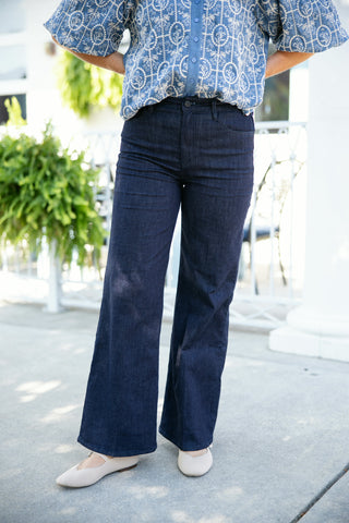 Lower-body front view highlighting high-waisted wide-leg flare jeans in a clean, dark indigo wash. The trousers feature a classic button-fly closure, front curved scoop pockets, and a full-length wide leg that drapes smoothly over cream pointed-toe flats, styled with a tucked-in blue embroidered puff-sleeve top on a sunny outdoor patio.
