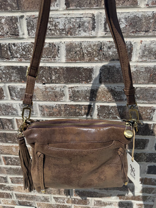 A front view of a vintage-inspired HOBO brand crossbody bag crafted from distressed brown leather with a subtle sheen, hanging outdoors against a textured brick wall in natural sunlight. The bag features a top zipper closure finished with a round antique brass branded pull, a convenient curved front slip pocket, and a matching decorative leather tassel detail on the left side. The design includes an adjustable, detachable leather strap secured by sturdy metal lobster clasps and O-rings, with the official wh