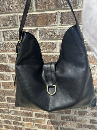 A front view of a slouchy black leather hobo-style shoulder bag hanging outdoors against a textured brick wall in natural sunlight. The bag features a classic relaxed silhouette with a deep U-shaped dipped topline. A prominent central leather strap detail with double topstitching drapes over the front, finished with a sleek, gold-tone metal D-ring hardware piece. The design includes a single adjustable flat leather shoulder strap secured with a gold-tone buckle on the left side, highlighting the smooth, nat