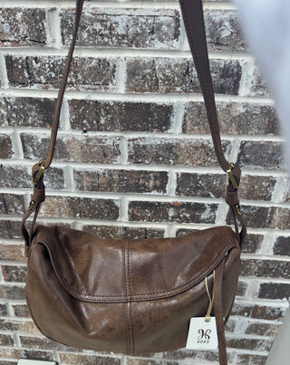 A front view of a vintage-inspired HOBO brand crossbody bag crafted from distressed, glazed brown leather with a subtle sheen, hanging outdoors against a textured brick wall in natural sunlight. The bag features a slouchy, soft-structured silhouette highlighted by a large curved front flap with double-stitched borders and a prominent vertical center seam. The design includes an adjustable flat leather shoulder strap secured with antique brass-tone rectangular buckles on both sides, with the official white H