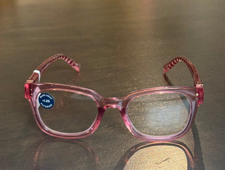 Front view of translucent pink square-frame reading glasses resting on a smooth metallic surface. A circular blue sticker on the left lens highlights its +1.25 magnification and blue light filtering capabilities, combining a stylish, glossy acetate finish with functional digital eye protection.