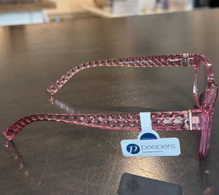 Side profile of translucent pink Peepers reading glasses, showcasing the intricate diamond-quilted architectural texture along the temple arms. The glasses rest on a metallic surface with an official "Peepers by Peeperspecs" brand tag hanging from the sculpted arm, highlighting the frame's premium detailing and glossy finish.