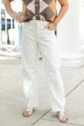 A detailed lower-body view highlighting the white straight-leg denim jeans, focusing on the intricate tonal white floral lace appliqués patched along the outer thighs and lower legs. Details the high-rise waist, classic five-pocket design, subtle raw fringe hem, and metallic gold mules on a sunlit paved sidewalk.