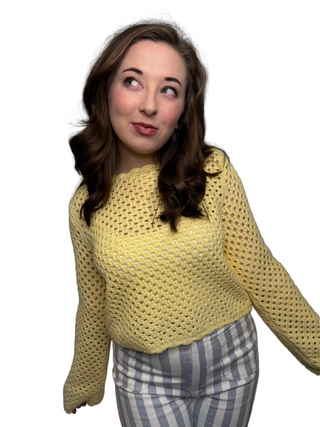 A woman wearing the Lemon Sherbert Top, a butter-colored open-knit crochet sweater with scallop trim on the crew neck and hem. She is looking to the side and smiling, styled with blue and white vertical striped trousers against a white curtain backdrop.